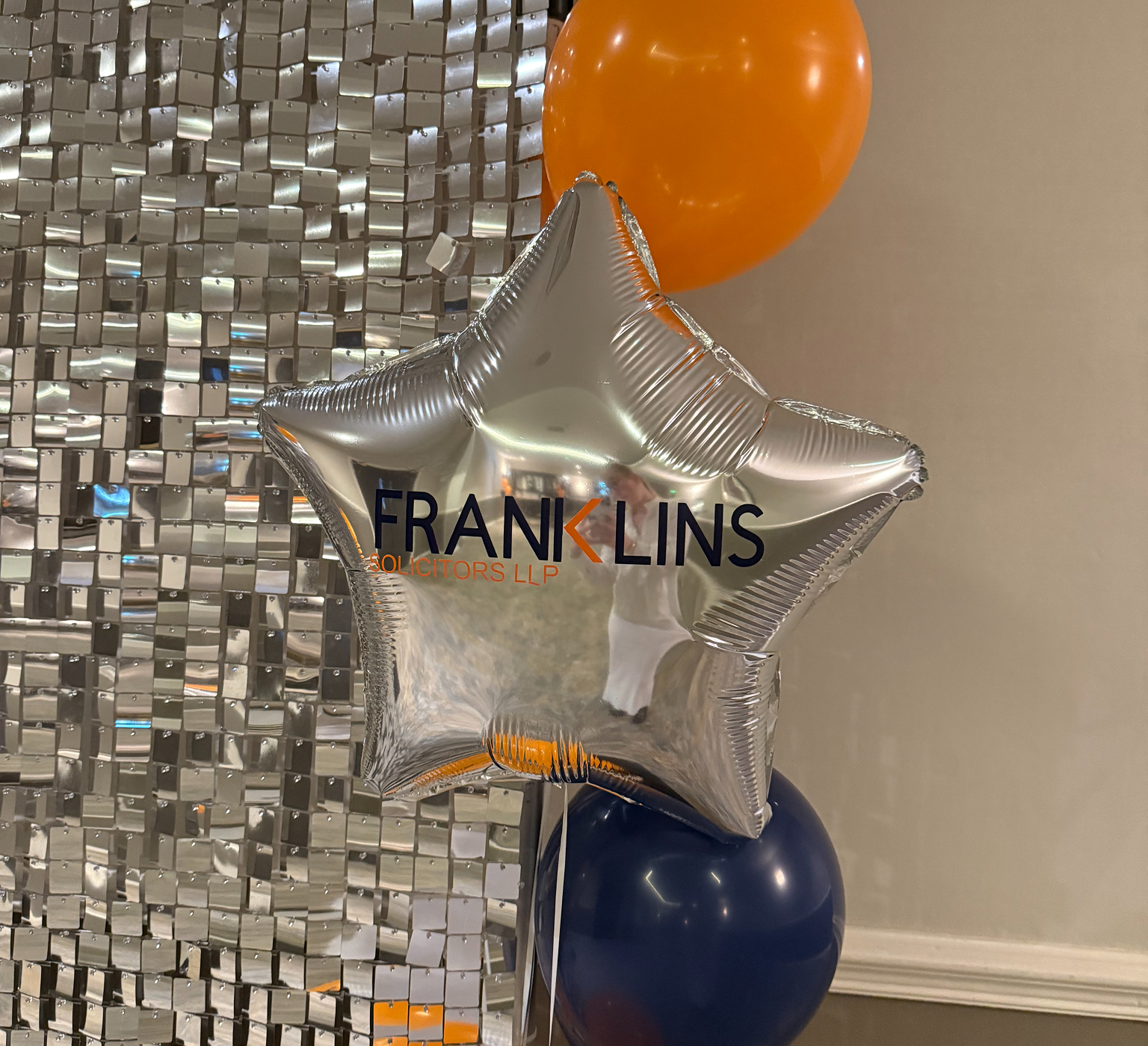 Silver star balloon printed with the Franklins Solicitors LLP logo, beside orange and navy balloons and a silver sequin wall
