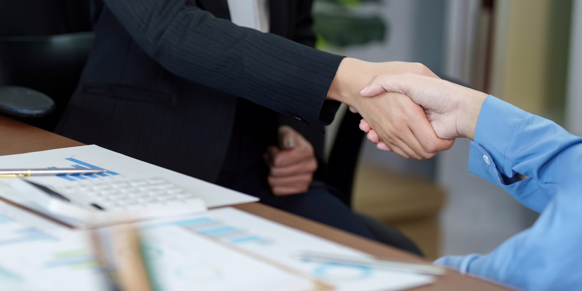 Two business professionals shaking hands across table with documents and charts