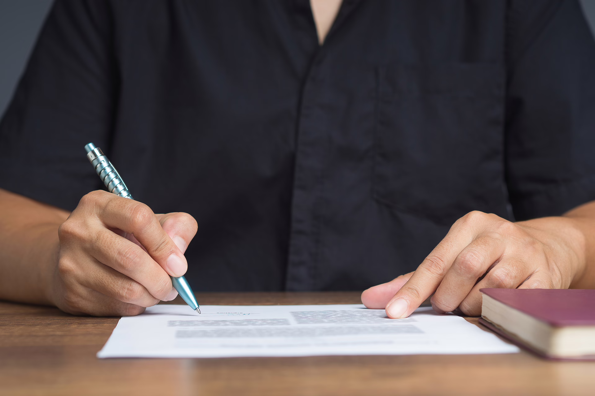 Person signing a printed document with a pen at a desk, with a closed book beside them.