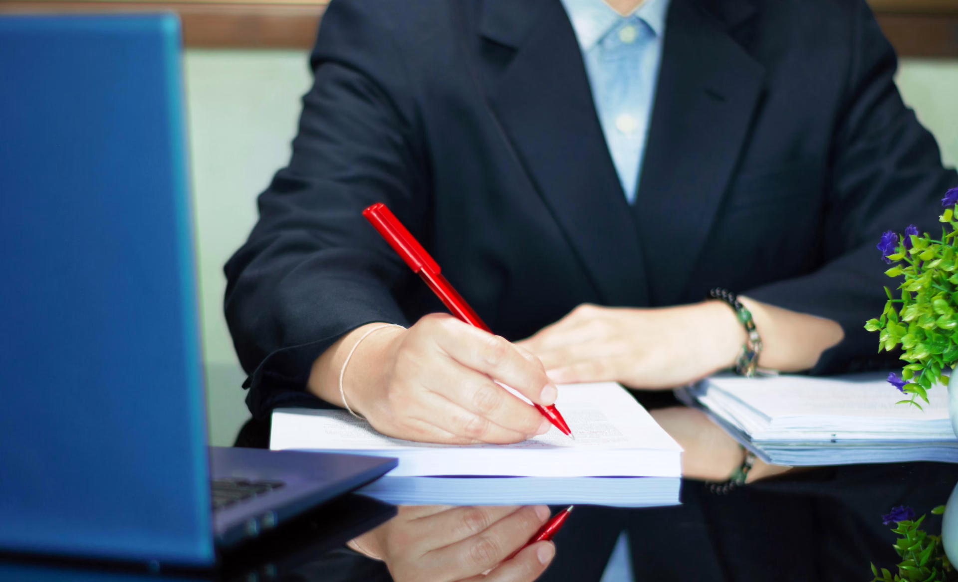 Person in a suit writing on a document with a red pen at a desk beside a laptop.