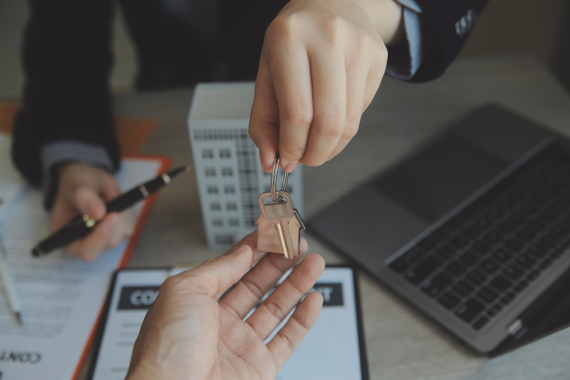 House keys being passed between two people across a desk with property paperwork and a laptop visible.