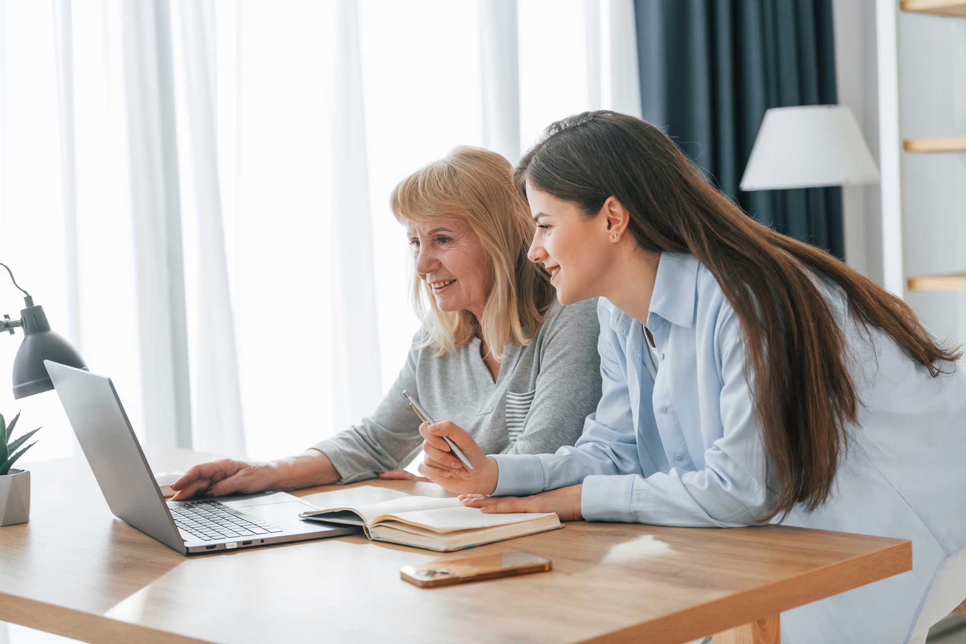 Two women sitting at a table looking at a laptop together in a bright room.